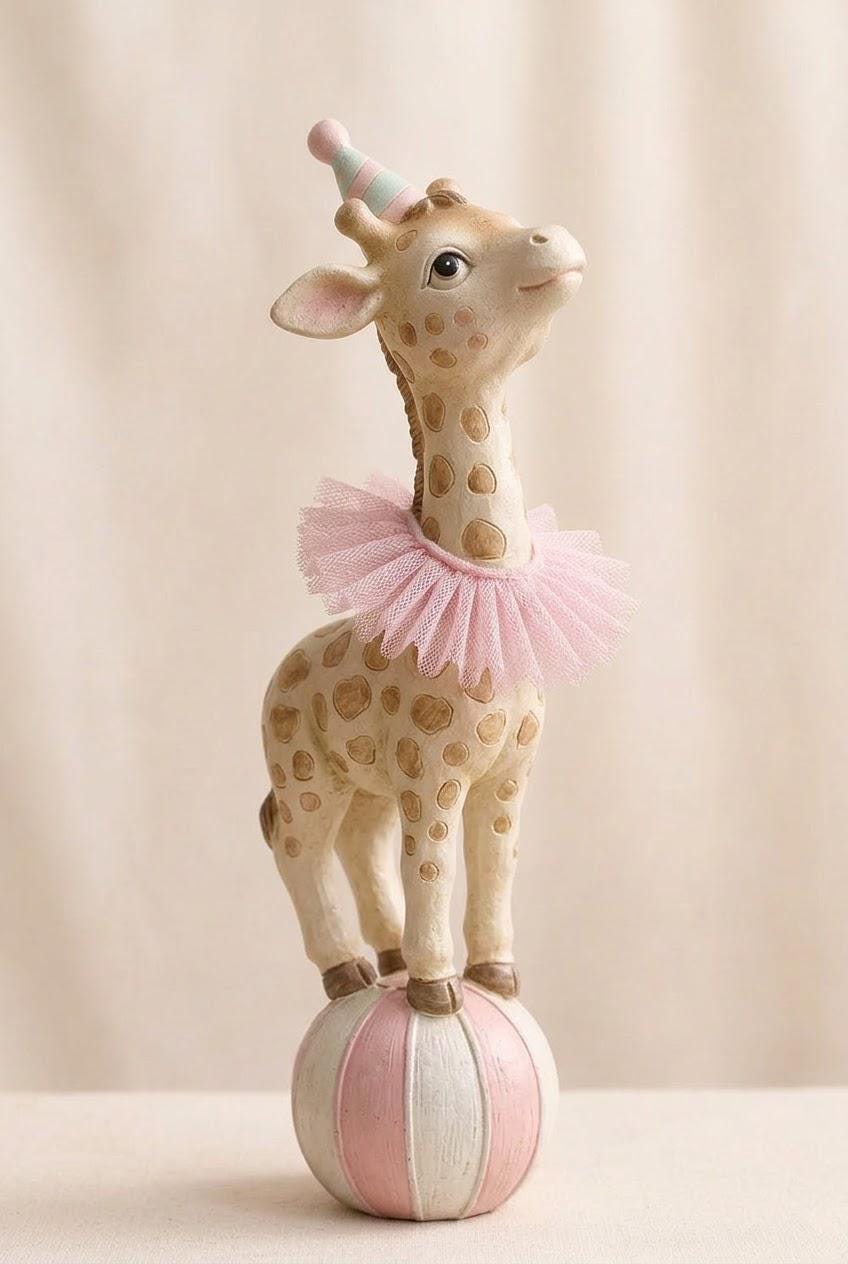 May include: A whimsical giraffe figurine with a party hat and pink tutu collar. The giraffe, with brown spots, stands on a striped pink and white ball. The figurine is a decorative item.